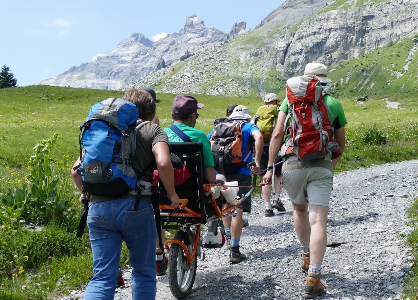 Sortie en joëlette sur un sentier ensoleillé face au massif du Mont-Blanc, entourée d'accompagnateurs souriants.