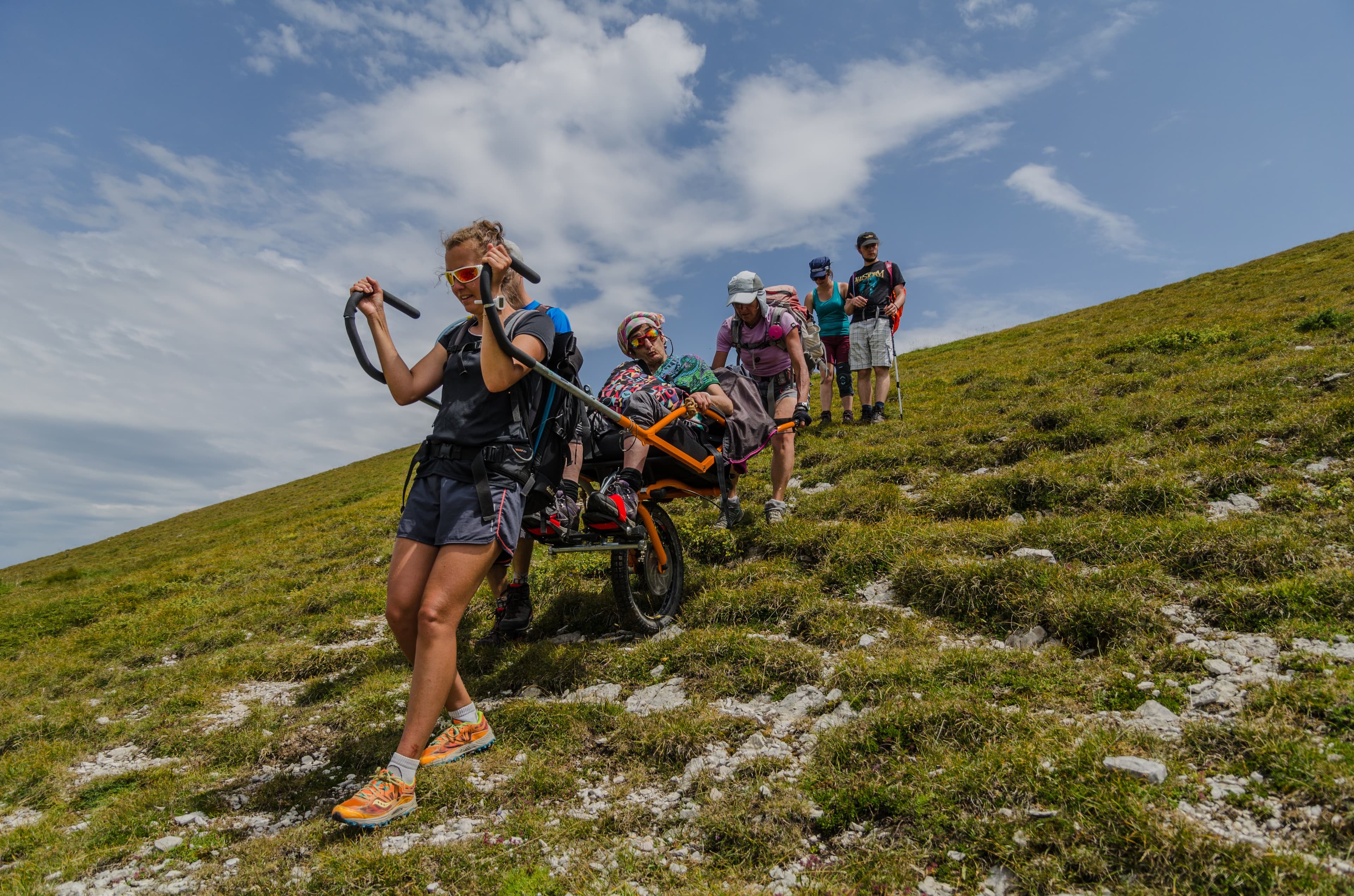 Sortie en joëlette sur une croupe herbeuse ensoleillée, l'accompagnatrice ouvre la marche et l'équipe suit sous un grand ciel bleu.