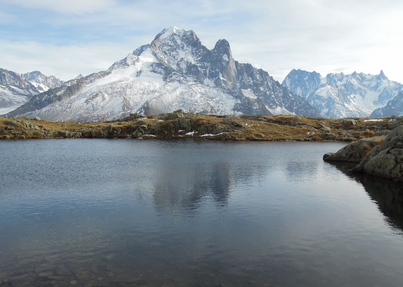L'Aiguille Verte et l'Aiguille du Dru enneigées se reflétant dans un lac d'altitude du massif du Mont-Blanc, prairie dorée à l'automne.